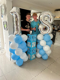Two people standing between large silver number '18' balloons in a kitchen.